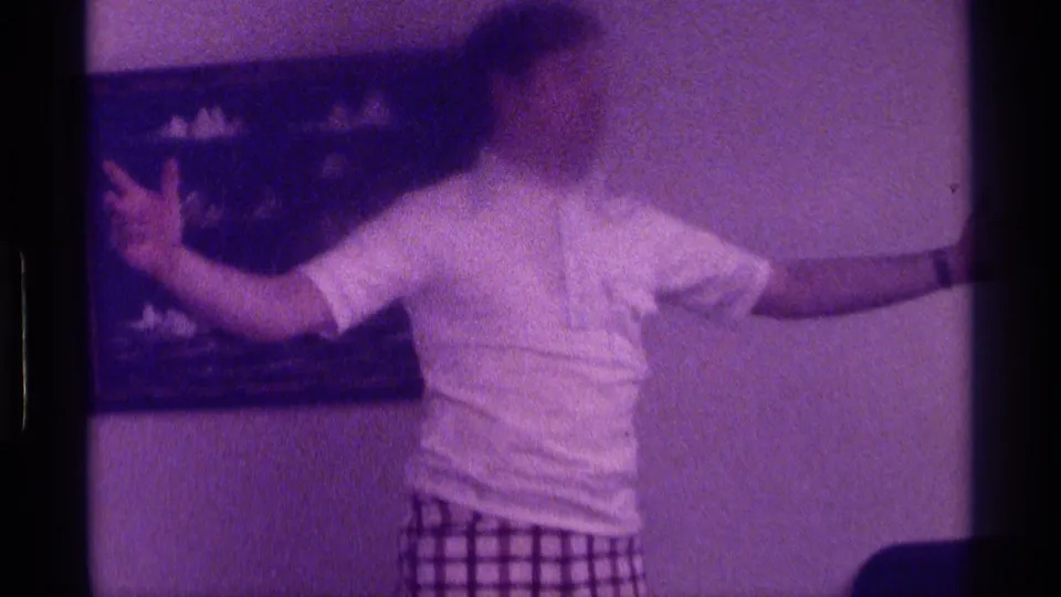 1976 Kentucky 8mm home-movie frame showing a person turned toward wall art with arms spread inside a private living room.