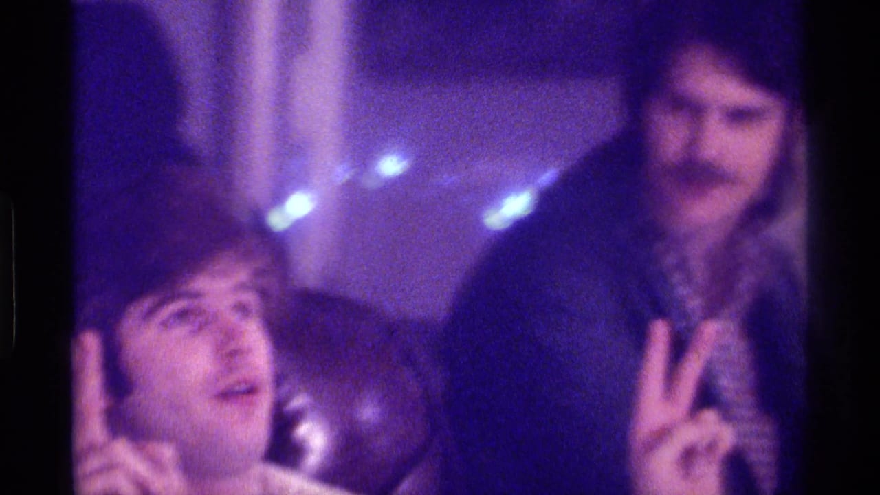 Two adult men in a dim room, one making a peace sign toward the camera.