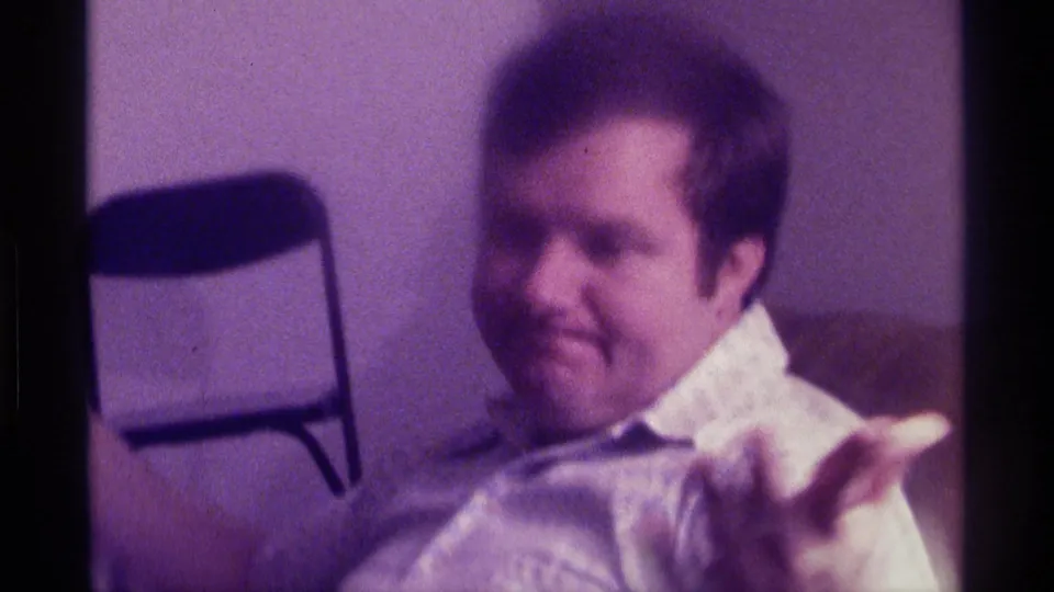 Kentucky 1976 home-movie frame of an adult man in a light shirt seated indoors, reacting casually to the camera.