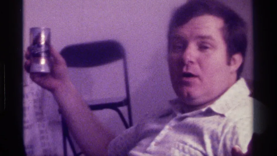 Kentucky 1976 home-movie frame of an adult man in a light shirt holding a can while seated in a private room.