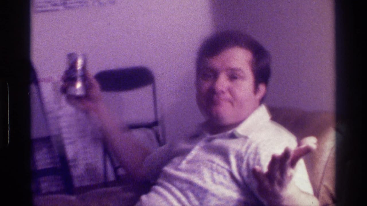 Man seated in a recliner raises a beer can and gestures toward the camera.