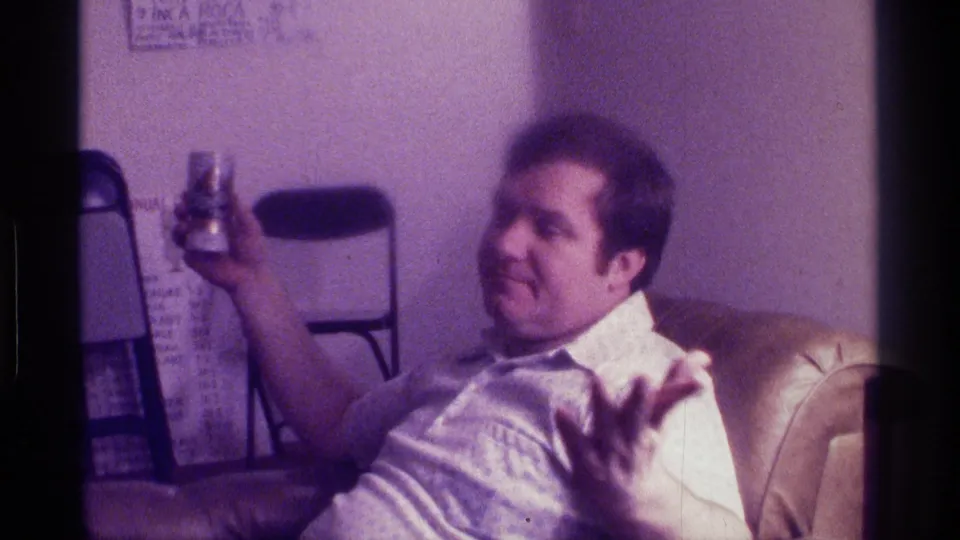 A soft, purple-tinted 8mm frame shows the man in a recliner looking to the side, with a beer can raised and folding chairs and wall writing behind him.