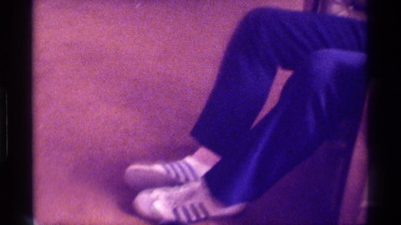In this 1976 Kentucky home-movie frame, a seated man’s lower legs show dark pants, striped socks, and white sneakers on carpet.