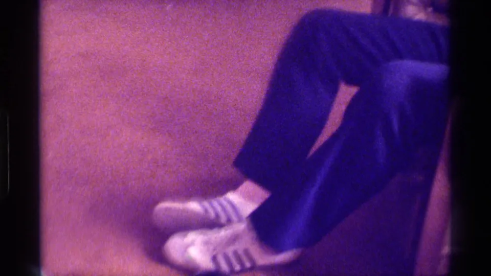 In this 1976 Kentucky home-movie frame, a seated man’s lower legs show dark pants, striped socks, and white sneakers on carpet.