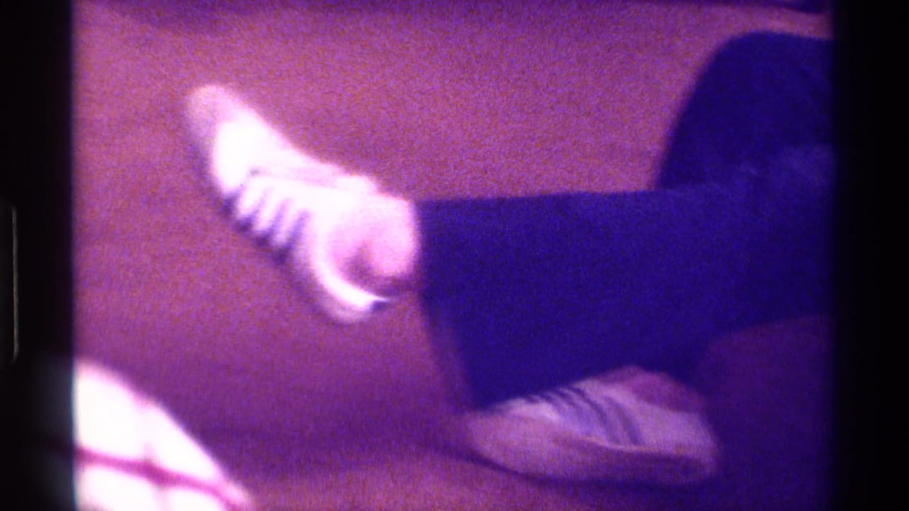 A close 1976 Kentucky frame shows one raised foot in a white striped sneaker, with the other shoe near the carpeted floor.