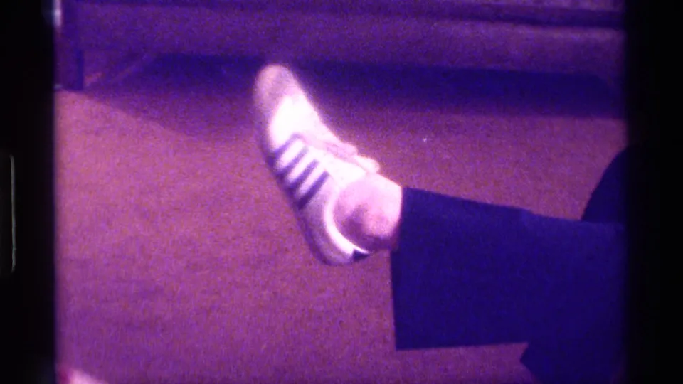 The home-movie frame isolates a lifted white striped sneaker, striped sock, and dark pant leg against the room’s carpet.