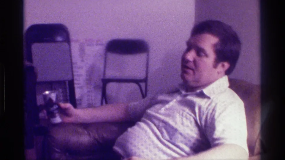 A man in a white shirt reclines in a 1976 Kentucky interior, with folding chairs and a carpeted room visible behind him.