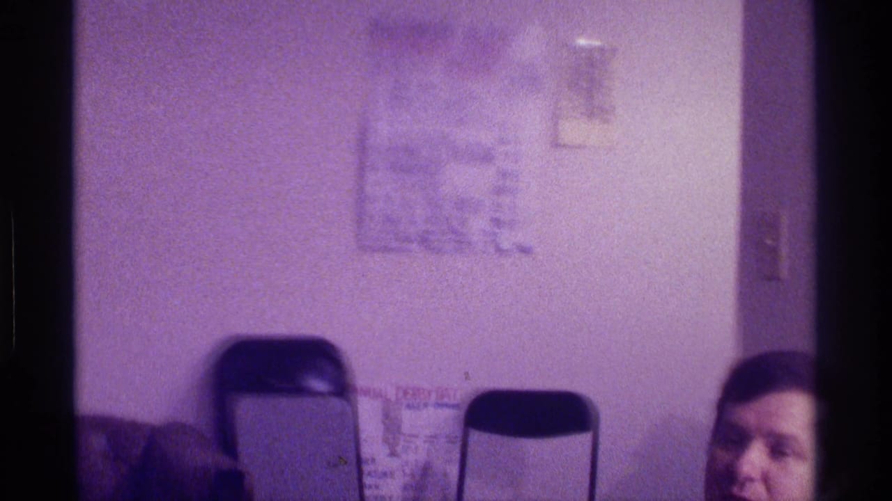 1976 Kentucky home-movie frame showing a dim living room with folding chairs, wall papers, and a man partly visible at the bottom edge.