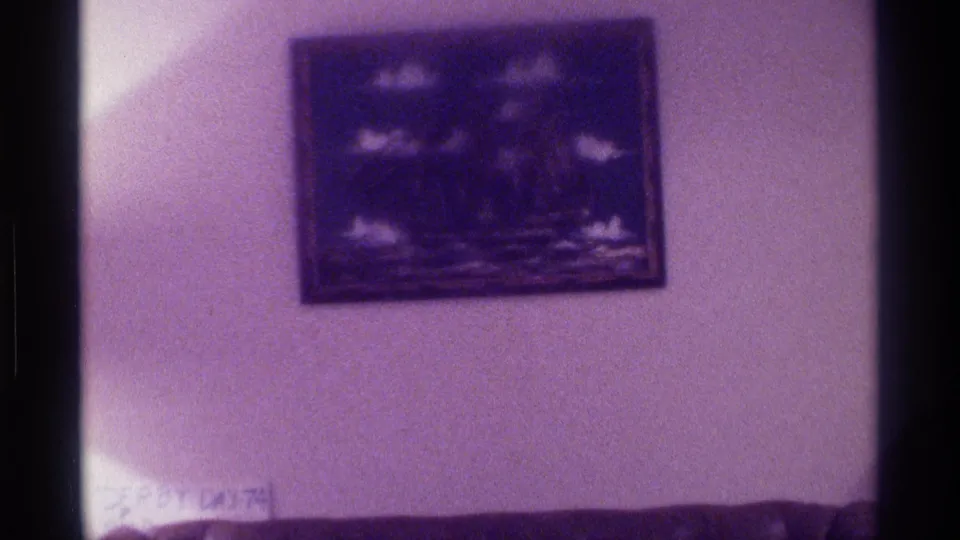 1976 home interior frame showing framed wall art over low furniture, with soft exposure and purple 8mm film color cast.