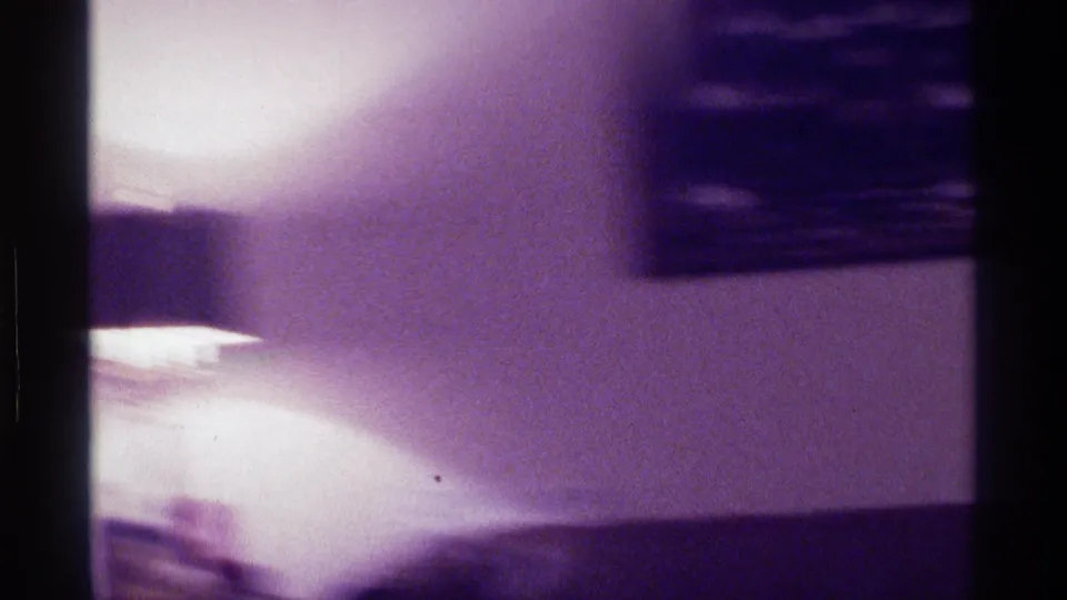 Blurred Kentucky home-movie frame with framed wall art, a couch or chair, and a bright overexposed patch during a handheld pan.