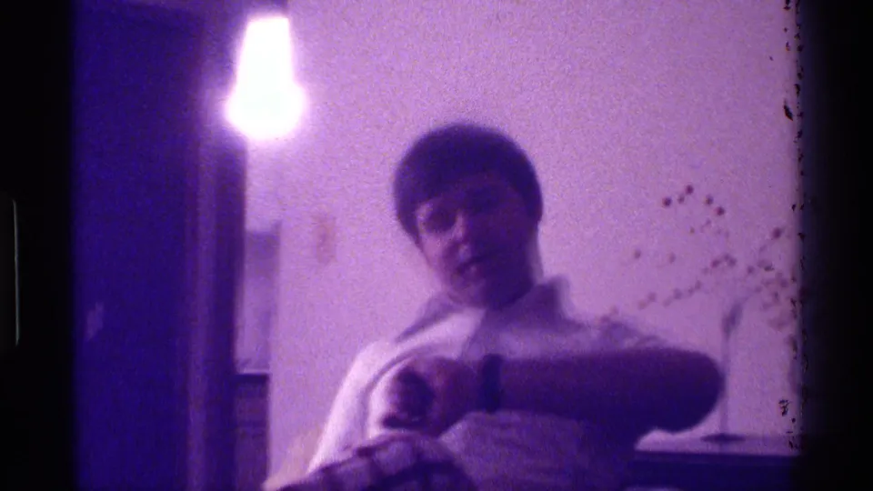 Young man seated on a couch gestures toward the camera in purple-tinted 1976 Kentucky home-movie footage.