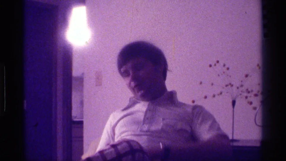 A young man turns to the side on a couch in a dim private interior, with 8mm grain and purple color shift.