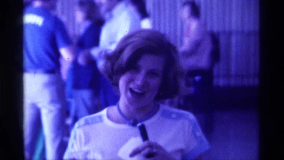 Smiling young woman holds a microphone at a Kentucky social gathering in 1976, with blurred people in the background.