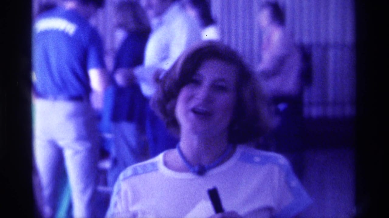 Woman in a light top smiles toward the camera in a dim indoor gathering.