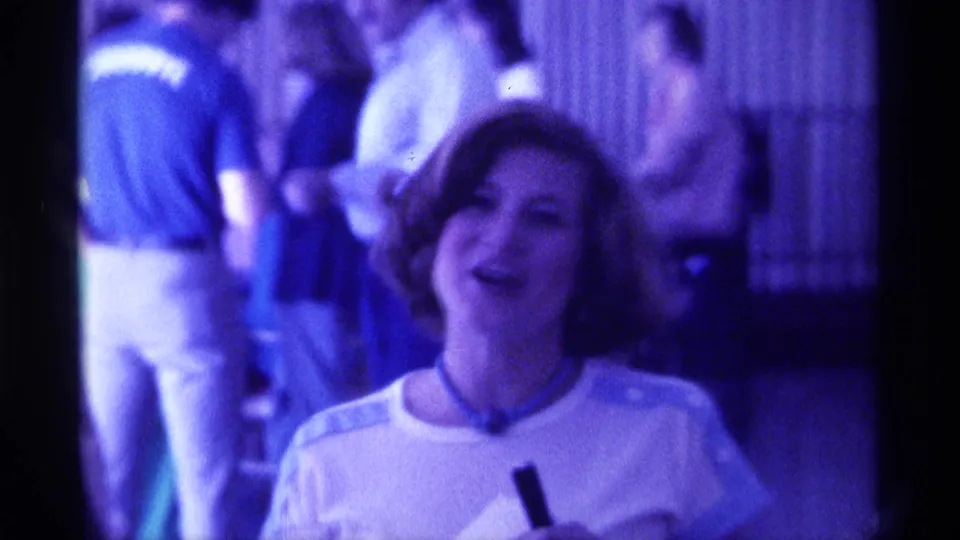 In 1976 Kentucky home-movie footage, a woman with short brown hair smiles toward the camera as people stand behind her indoors.