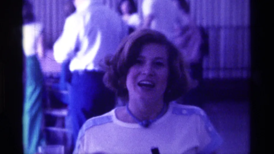 At the end of the clip, the woman faces the camera and appears to speak or laugh while adults remain visible in the indoor background.