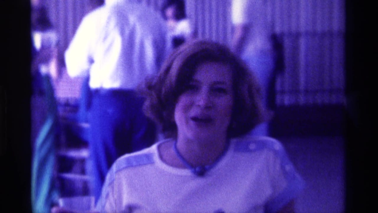 The woman smiles mid-speech in a purple-lit Kentucky indoor setting, with indistinct figures and vertical blinds behind her.