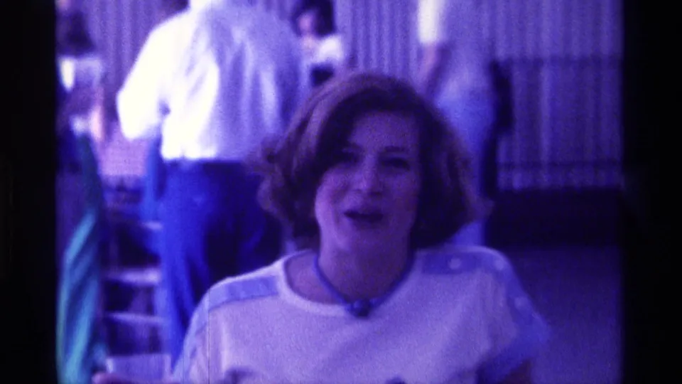 The woman smiles mid-speech in a purple-lit Kentucky indoor setting, with indistinct figures and vertical blinds behind her.