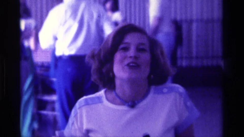 A young woman continues speaking in grainy 8mm footage, her face softly focused under strong purple indoor light.