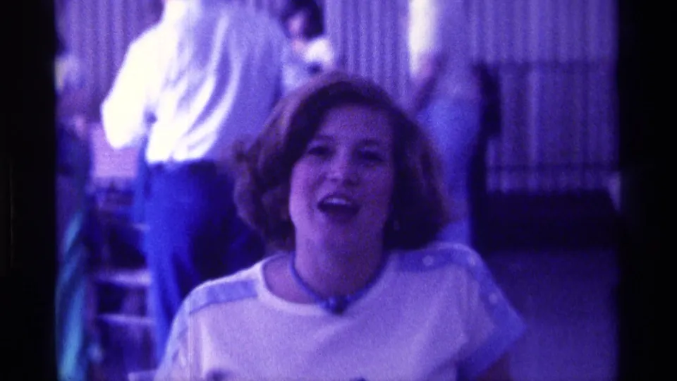 The woman appears mid-sentence in a Kentucky home-movie close-up, with dim figures and vertical blinds in the background.