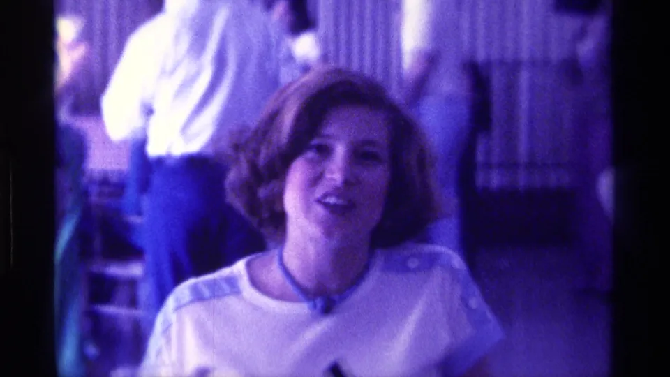 The woman appears to speak toward the camera, with standing adults behind her in a purple-tinted 8mm indoor scene.