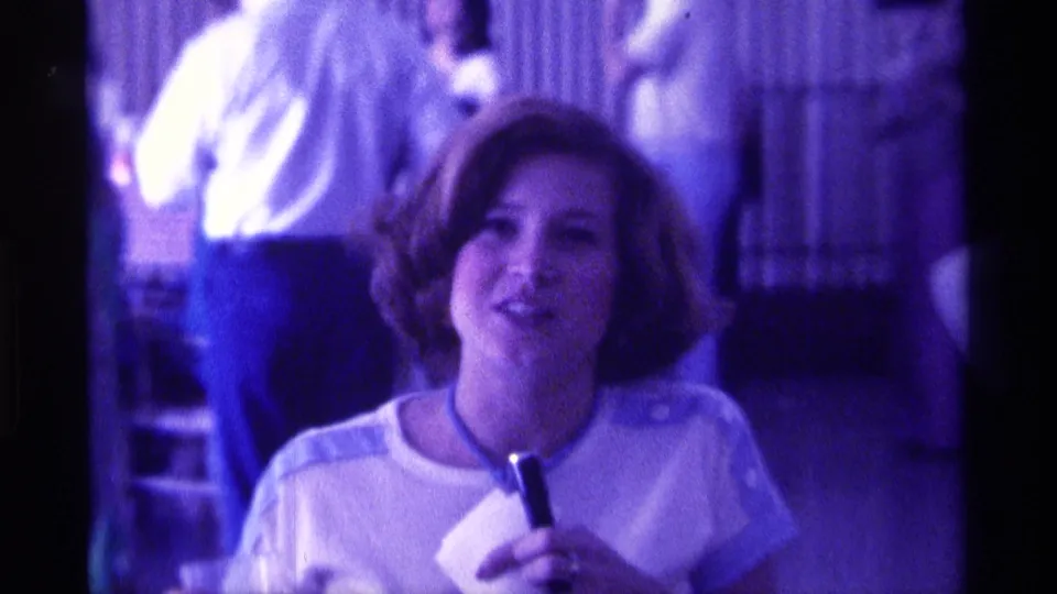 The woman holds a small microphone and light-colored paper while facing the camera in 1976 Kentucky home-movie footage.