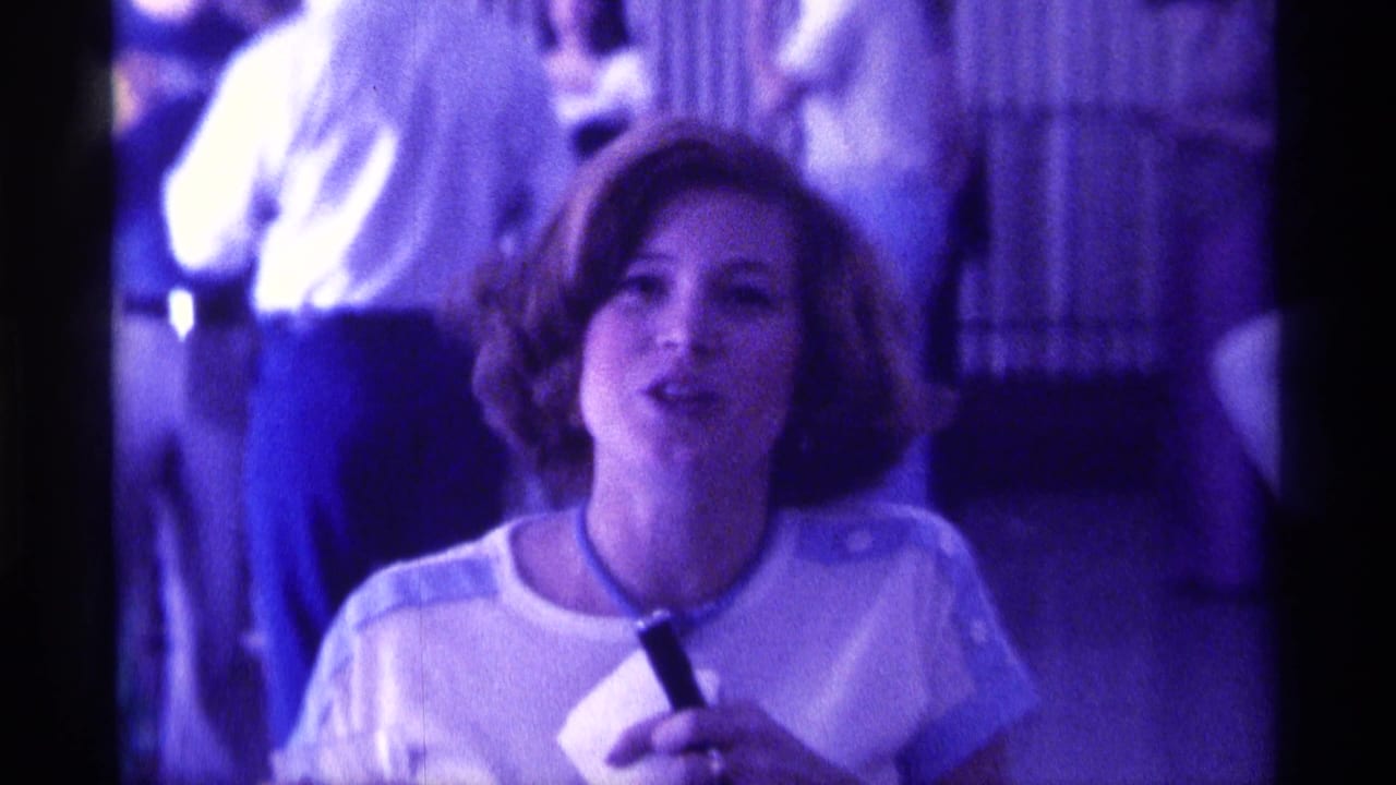 In 1976 Kentucky home-movie footage, a woman in a light top holds a microphone as guests stand behind her indoors.
