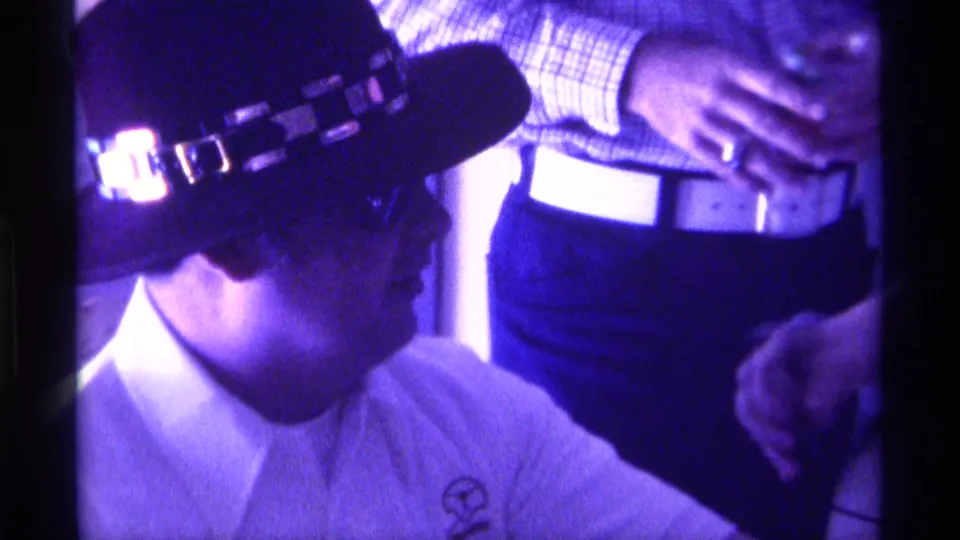 A seated man in a wide-brimmed hat is shown near adults standing close by at the 1976 Kentucky indoor gathering.