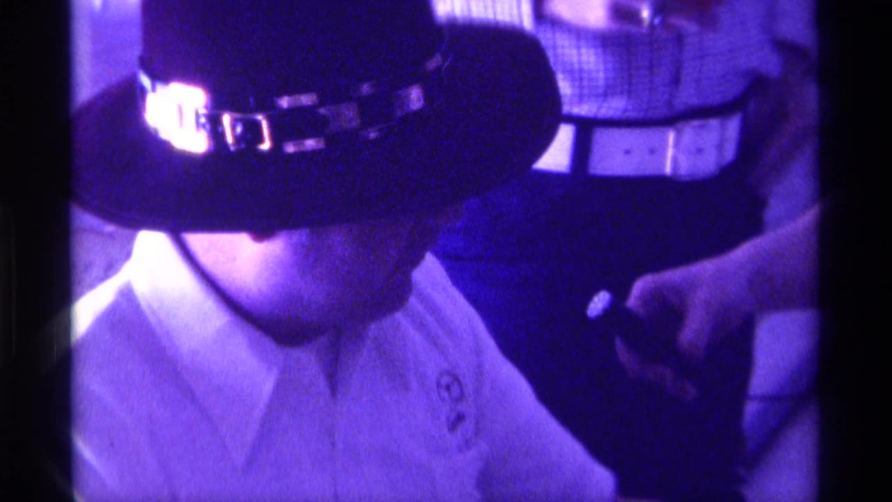Seated man in a dark decorative hat and white collared shirt beside standing figures indoors.