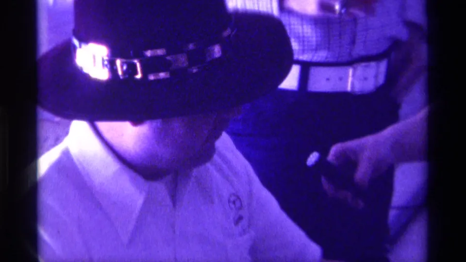 1976 Kentucky 8mm frame of a seated man in a dark hat and white collared shirt, with nearby people partly visible in purple-tinted indoor light.