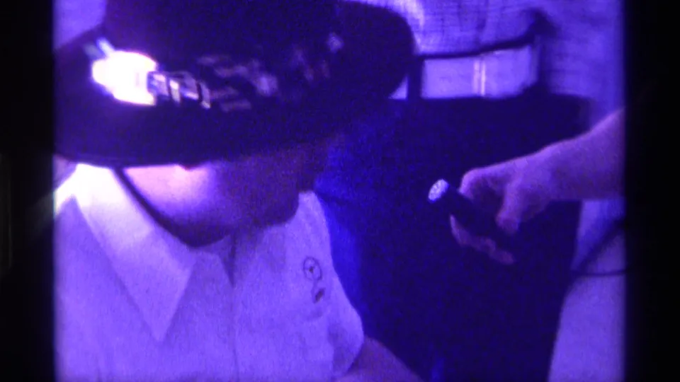 1976 Kentucky home-movie frame showing a seated man in a dark decorative hat as a nearby hand holds a small object near his shoulder.