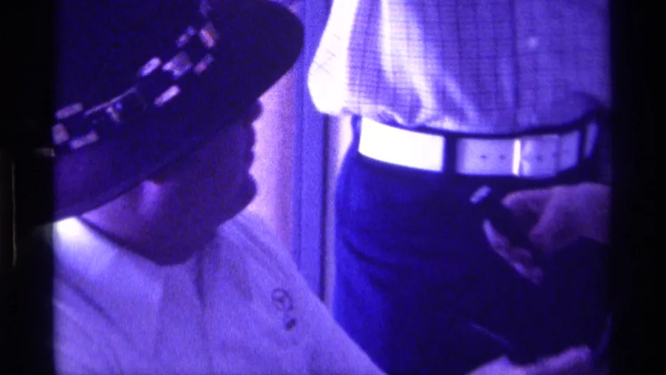 Side view from 1976 Kentucky 8mm footage of the seated man in a dark hat, with a checkered shirt and white belt visible behind him.