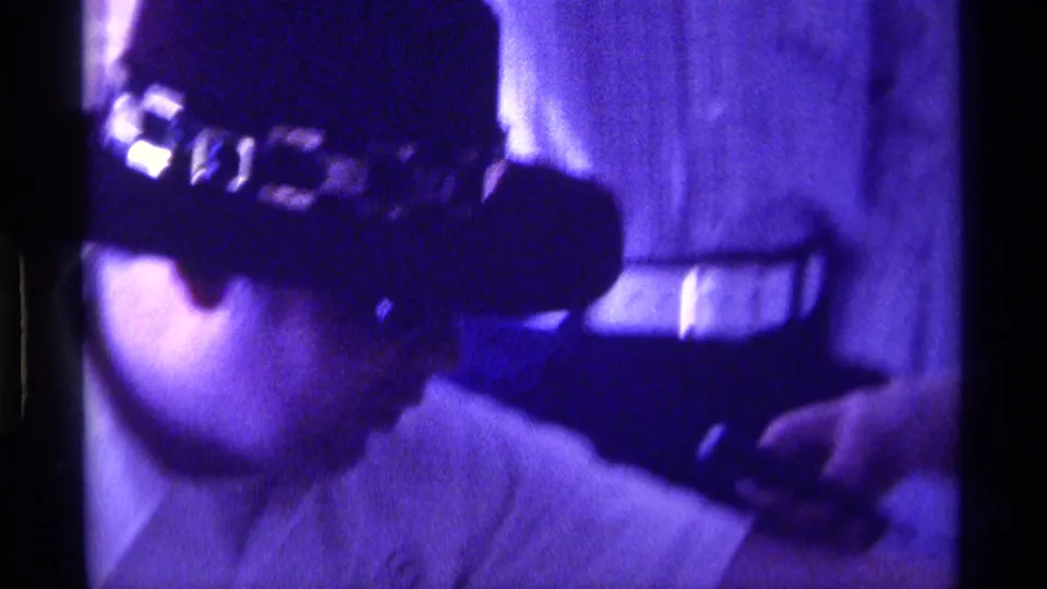 Purple-tinted 1976 Kentucky 8mm frame showing the man in the dark decorated hat from the side, with a hand and small object near him.