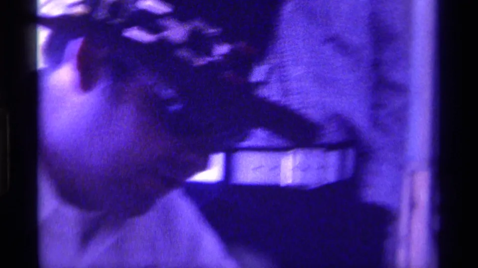 Purple-tinted home-movie frame of a man in a dark cowboy hat seen in soft profile inside a private Kentucky interior.