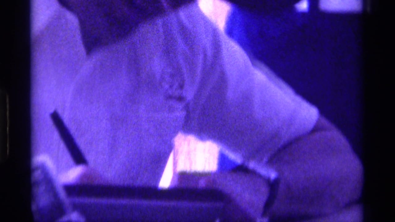 Man in a light shirt and dark hat leans over a tabletop in purple low light.