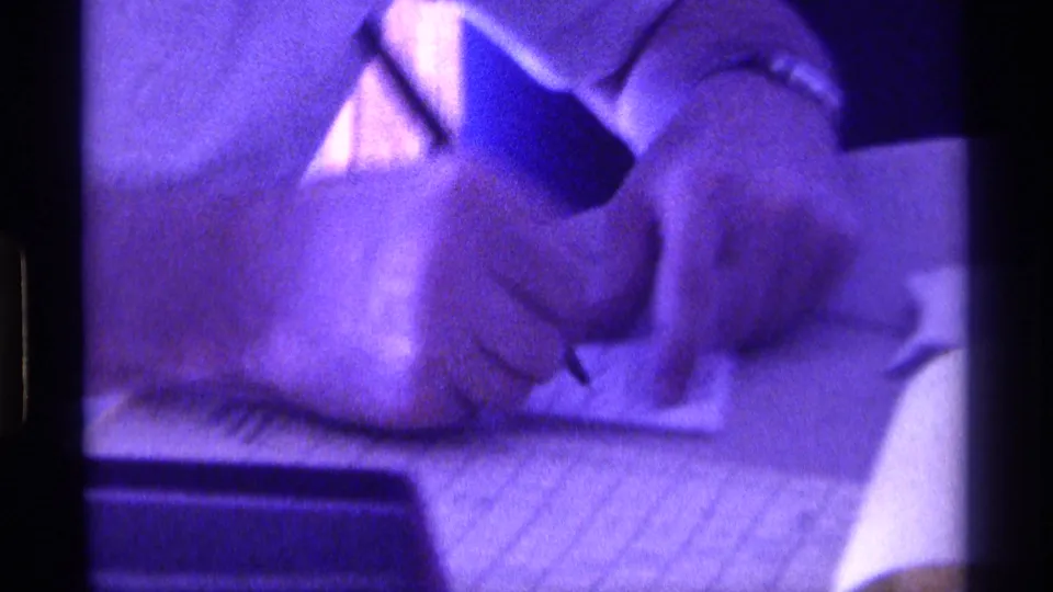 Close 8mm view of hands writing with a pen on a notepad, tinted purple by the low-light exposure.