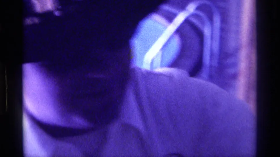 1976 Kentucky frame showing the man bending forward, with a dark hat and checkered band visible in purple-tinted low light.