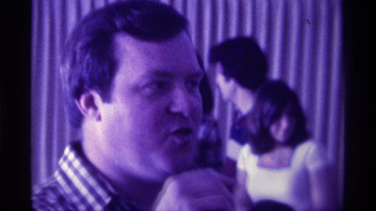 Man in a checkered shirt speaks near guests under purple indoor lighting.