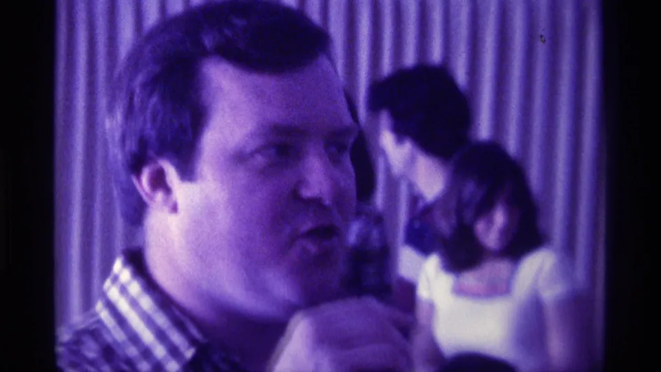 1976 Kentucky 8mm frame of a man in a checkered shirt speaking at an indoor party with guests blurred behind him.