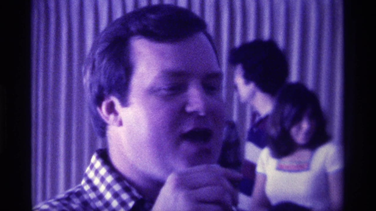 Close 1976 Kentucky party frame showing the speaker in profile-front view, with blurred guests and curtains in purple light.
