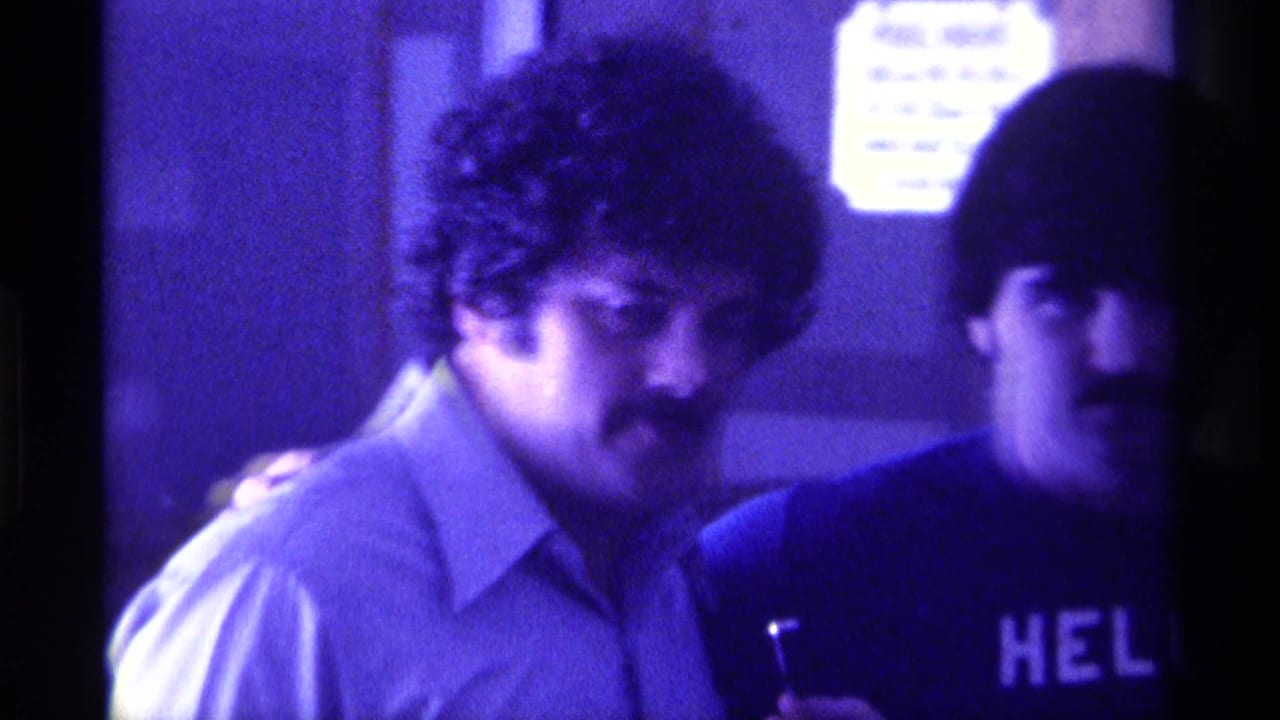 1976 Kentucky party frame with two men in purple-tinted light, one holding a glass and another wearing a shirt with partial lettering.