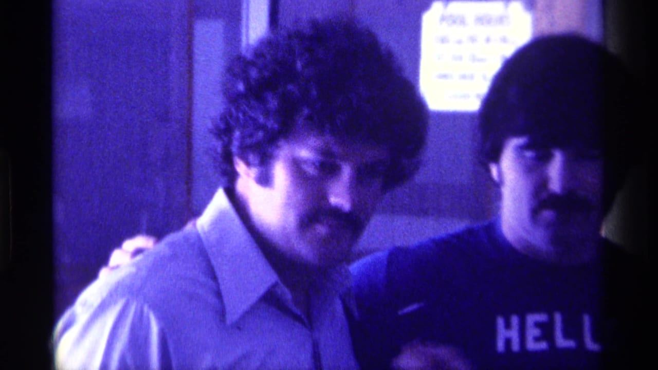 Two mustached men stand indoors, one in a light collared shirt and one in a dark HELLO shirt.