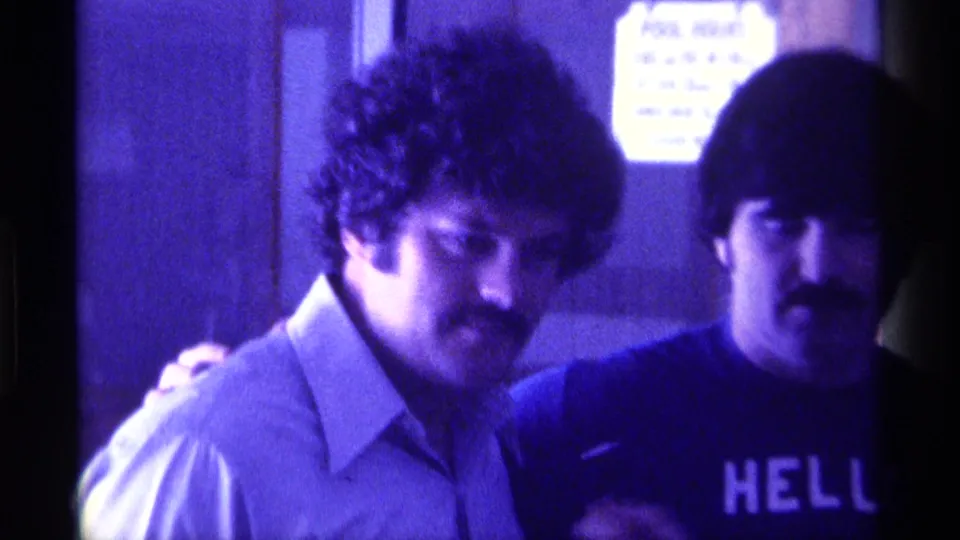 1976 Kentucky 8mm frame of two adult men indoors, one facing the camera in a light collared shirt and another beside him in a dark HELLO shirt.