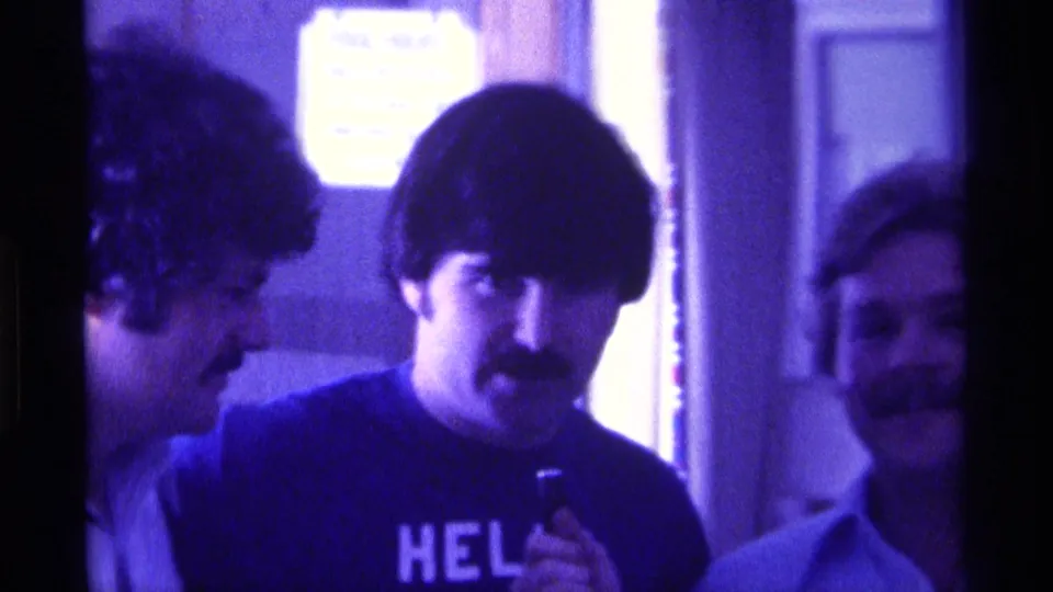 1976 Kentucky home-movie frame showing men in conversation indoors, including a man in a dark HELLO shirt holding a small microphone.