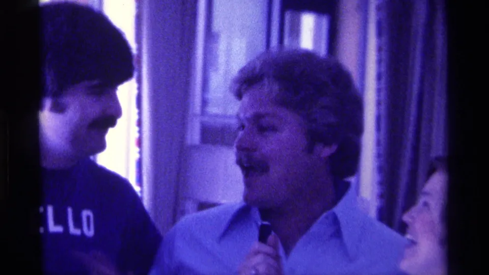 1976 Kentucky 8mm frame of a smiling man in a light collared shirt holding a microphone during an indoor home-movie conversation.