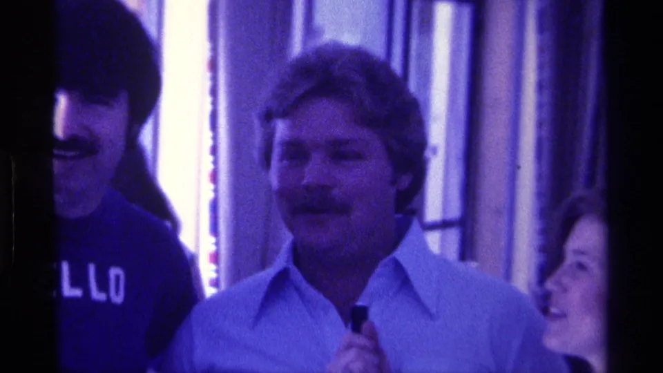 1976 Kentucky home-movie frame with a smiling mustached man holding a microphone as another man in a dark HELLO shirt stands beside him.