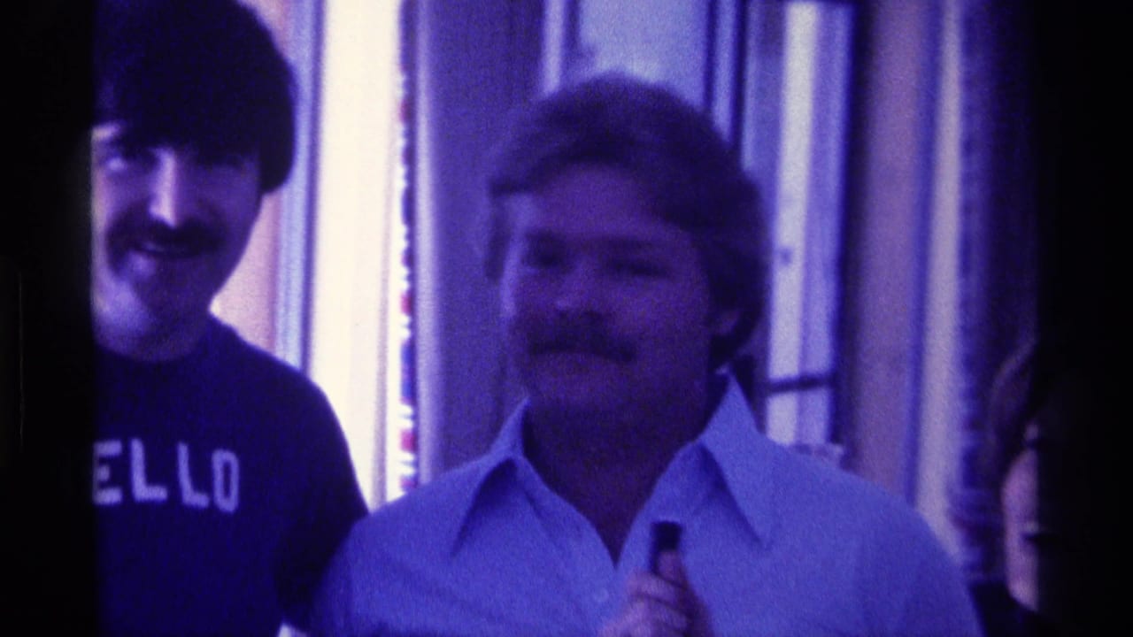 1976 Kentucky 8mm frame of two adult men smiling indoors, with one dark shirt showing the word HELLO.
