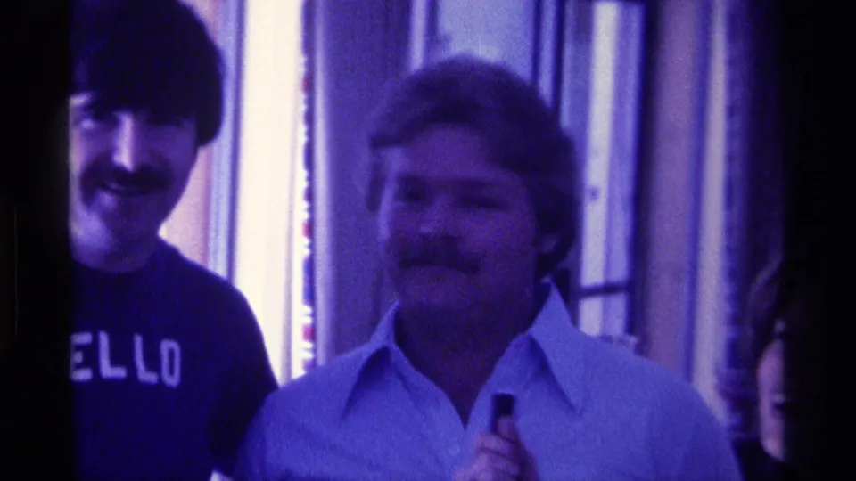 1976 Kentucky 8mm frame of two adult men smiling indoors, with one dark shirt showing the word HELLO.