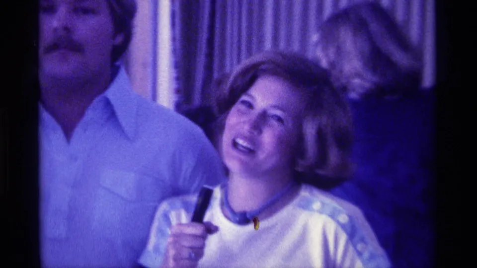 1976 Kentucky home-movie frame showing a woman laughing with a microphone as an adult man stands nearby indoors.