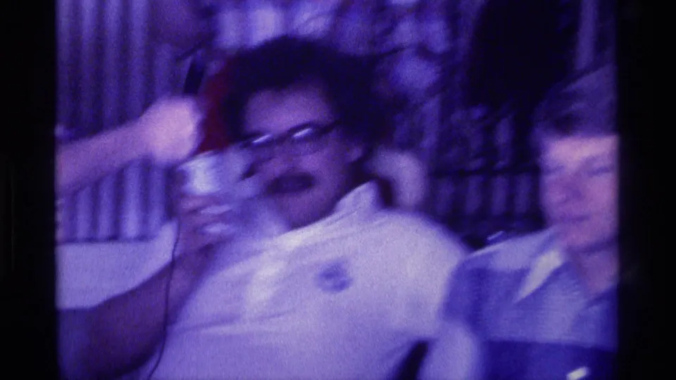 1976 Kentucky 8mm frame of a mustached man with glasses laughing or reacting as people gather around him indoors.